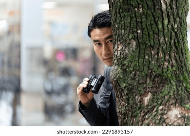 A man hiding in a tree and keeping a lookout with binoculars in the city.