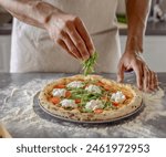 A man from a high angle view is placing arugula on baked pizza dough.