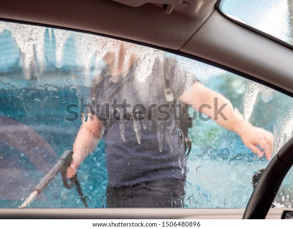 Man\
with the high powered hose in his hand washes his car at a\
self-serve car wash. View from the driver\'s\
seat.