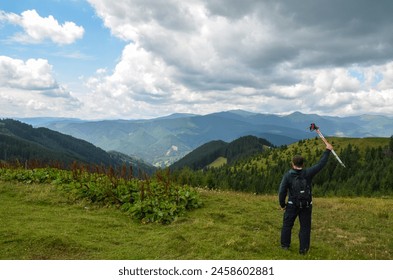 Man hiker with a backpack stands on a grassy hill raising in the hand trekking poles admires the panorama of undulating green mountains, the slopes of which are covered with dense forests. Carpathians - Powered by Shutterstock