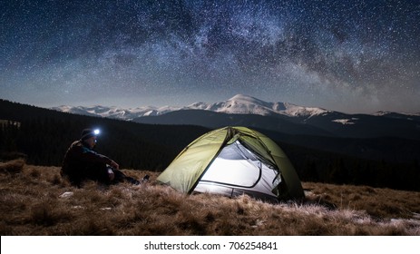 Man Hiker Enjoying Night Scene In His Camping In The Mountains At Night. Man With A Headlamp Sitting Near Tourist Tent Under Beautiful Night Sky Full Of Stars And Milky Way.