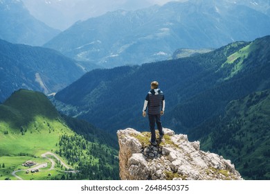 Man hiker standing on a cliff is enjoying the breathtaking view of the surrounding mountain range, green valleys, and winding road in the Italian Dolomites under a dramatic cloudy sky. Summer in Alps