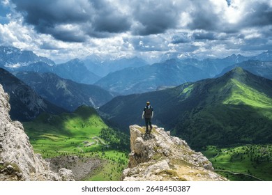 Man hiker standing on a cliff is enjoying the breathtaking view of the surrounding mountain range, green valleys, and winding road in the Italian Dolomites under a dramatic cloudy sky. Summer in Alps