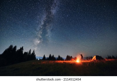 Man hiker standing on grassy hill near campfire and pointing finger at beautiful night starry sky with Milky Way under mountain valley. Concept of hiking, night camping and astrology. - Powered by Shutterstock