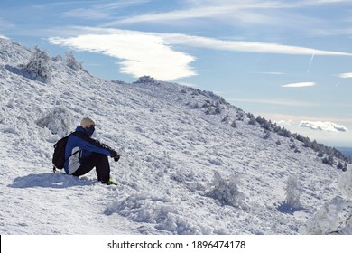 A Man With A Hiking Backpack Is Sitting In The Snow Mountain Contemplating A Panoramic View Of The Snowy Landscape On A Sunny Winter Day. He Is Wearing A Mask. Sport And Active Nature Life Concept.