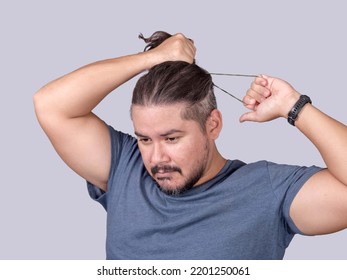 A Man In His 30s Ties His Long Hair Into A Ponytail With An Elastic Band. A Guy In A Gray Shirt Fixing His Messy Hair. Isolated On A Gray Background.