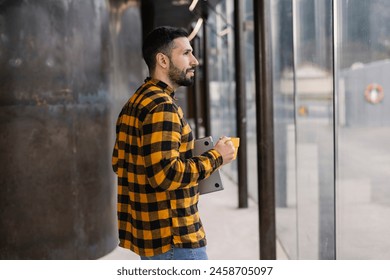 Man in his 30s, wearing a plaid shirt and jeans, walks thoughtfully with a coffee cup and tablet in a cityscape. - Powered by Shutterstock