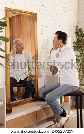 Man and his elder self reflected in mirror, both touching their faces looking at age-old changes of skin. Concept of human age, present, past and future, age, lifestyle, generation.