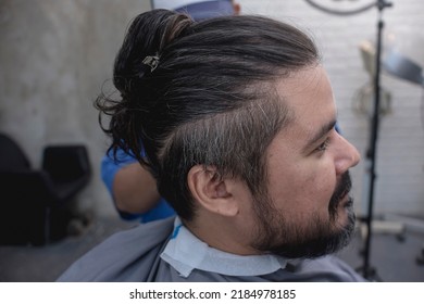 A man in his late 30s at the barbershop. His long hair tied up in a man bun in preparation for a haircut. Covered a barber cape tied around the neck lined with paper strip. - Powered by Shutterstock