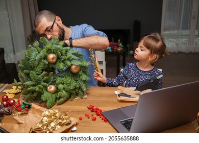 Man And His Little Daughter Learning To Decorate A Christmas Wreath Watching Lesson Online. Family Making Christmas Wreath. New Year Holiday Decoration. Christmas Tree Wreath Decoration DIY.