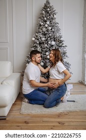 A man and his pregnant wife pose against the background of a Christmas tree - Shutterstock ID 2210226755
