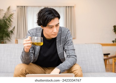 A man in his thirties who drinks tea while looking at a laptop in the living room