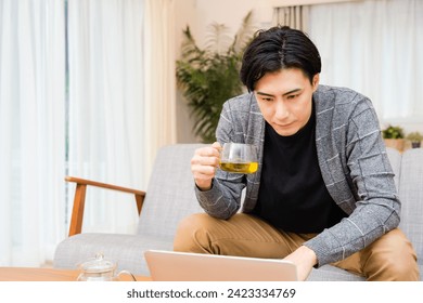 A man in his thirties who drinks tea while looking at a laptop in the living room
