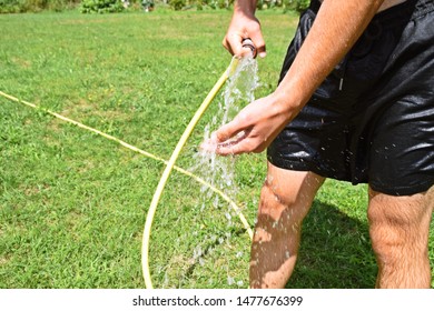 A Man In His Twenties In A Black Bathing Suit Refreshes Himself With A Garden Hose - Focus On The Legs Of The Man In A Green Meadow