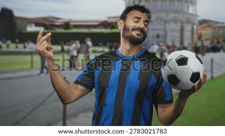 Man hispanic middle age with beard in blue striped soccer uniform holding a soccer ball in one hand and making a pinched fingers gesture at building lawn and walkway; confidence swagger.