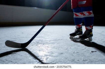 Man Hockey Player In Sports Uniform And Skates Standing On Ice Arena With Stick In His Hands. An Athlete Hones His Skills In Playing Hockey On Ice Of Stadium In Dark With Backlight. Legs Close Up.