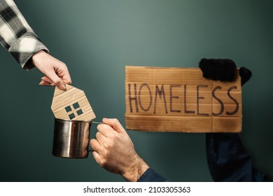 A man hold a cardboard sign with the inscription HOMELESS and steel cup. Woman put a carved mini house in mug. Dark greeny background. Hands close-up. The concept of helping vagabonds.