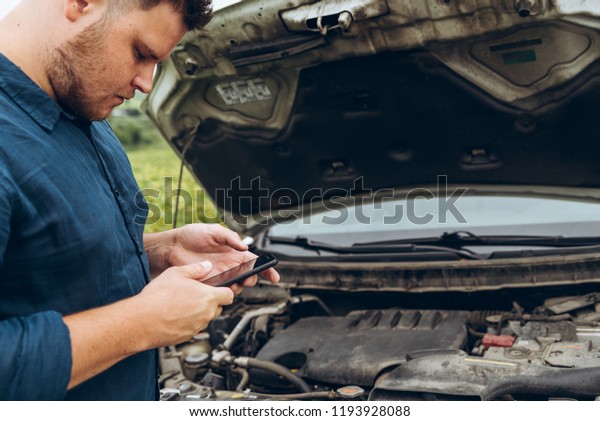man hold phone in hand. opened hood on background.\
broken car