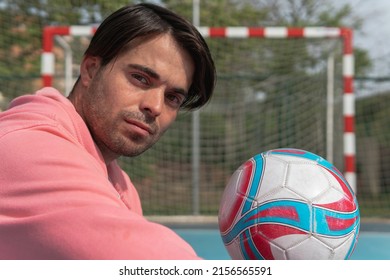 Man Holding The Ball In His Hand And Looking At The Camera On A Blue Soccer Pitch. Close Up Shot Of A Young Boy In A Pink Sweatshirt Lifting A Red, White And Blue Ball With The Goal In The Background.