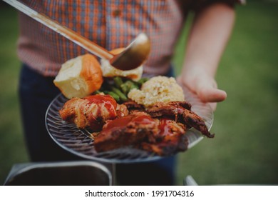 Man Holding A BBQ Plate Whole Pouring BBQ Sauce All Over Food. Backyard Texas BBQ On A Summer Day. 
