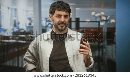 Man holding beer bottle with hand, wearing brown shirt and white overshirt, standing in an airport seating area; casual relaxation.