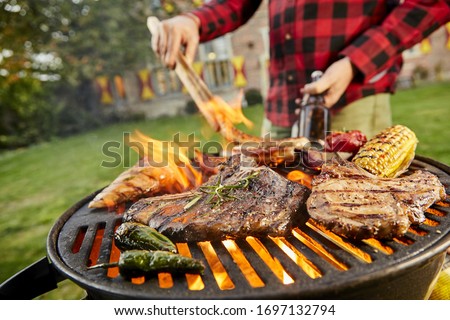 Man holding a beer grilling meat on a BBQ outdoors in his garden turning the sausages with tongs in a close up view on the beef steaks, corncob, chili peppers and chicken breast