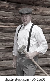 A Man Holding A Big Sledge Hammer By A Log Cabin