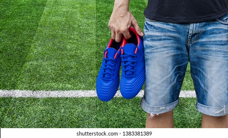 Man Holding Blue Football Boots In His Hand On Soccer Field.