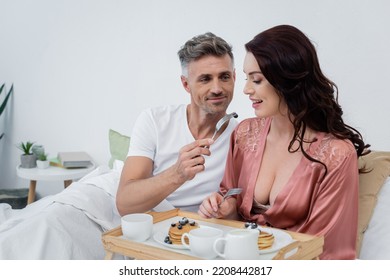 Man Holding Blueberry On Fork Near Wife In Silk Robe And Breakfast On Tray On Bed