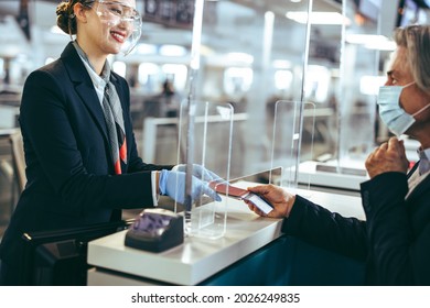 Man Holding Boarding Pass And Passport At Airline Check-in Desk At International Airport During Pandemic. Airport Employee In Face Shield And Passenger With Face Mask.