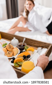 Man Holding Breakfast Tray With Orange Juice And Fruits Near Girlfriend On Hotel Bed On Blurred Background