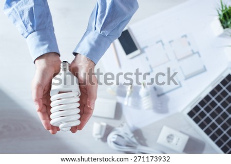 Man holding a CFL energy saving lamp, solar panel and house project on background, top view