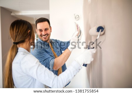 Man holding color palette and showing it to a woman while she painting wall with roller.