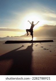 Man Holding Crane Stance while Balancing on a Log on the Beach Backlit by The Sun