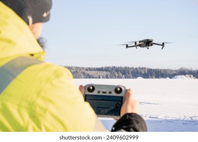 A man holding a drone remote controller in his hand and a drone flying in the blue sky. Winter season photo with snow field and forest on the background.