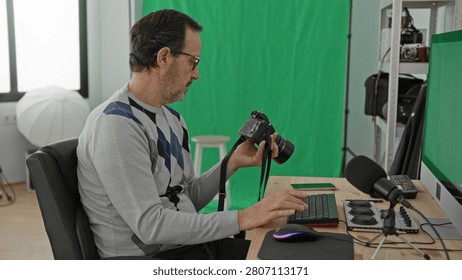 Man holding dslr camera while typing on keyboard at a desk in studio, inspecting lens and camera strap beside microphone and green screen; concentration.