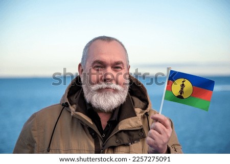 Man holding FLNKS New Caledonia flag. 
Portrait of older man with a national flag. 
Visit Travel to New Caledonia. Older man 50 55 60 years old 
with gray beard outdoors traveling in winter.