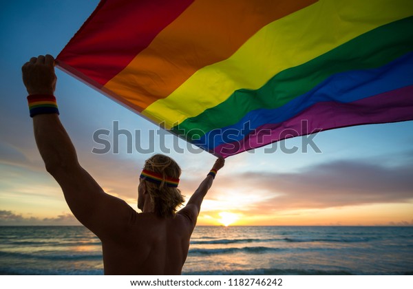 日の出の海岸で羽ばたく虹のゲイのプライド旗を持つ男性 の写真素材 今すぐ編集