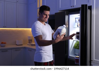 Man Holding Gallon Bottle Of Milk Near Refrigerator In Kitchen At Night