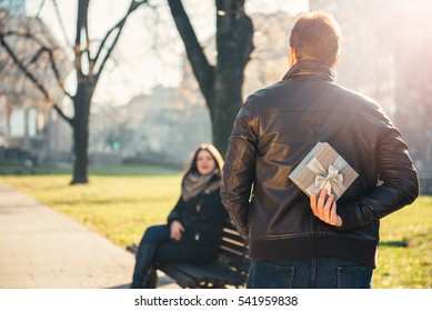 Man Holding A Gift Behind Back, Woman Siting On The Park Bench In The Background