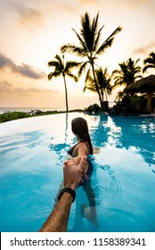 Man Holding Hand of Young Beautiful Woman in Clear Blue Infinity Pool in Tropical Island Paradise with Palm Trees and Colorful Sunset Background