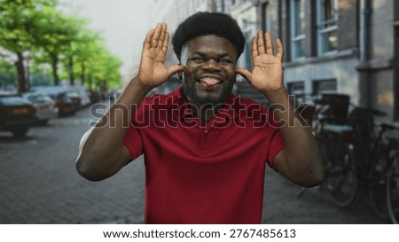 Man holding hands to ears while smiling broadly on a street beside parked bicycles and cars in a red polo; playful joy.