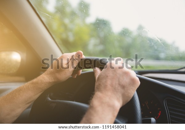 Man is holding hands on\
the car\'s steering wheel. Raindrops on the windscreen. Guy is\
traveling in the bad weather. Dangerous conditions. Lifestyle\
concept.