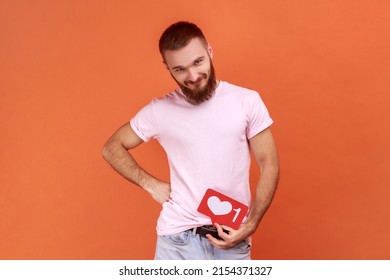 Man Holding Heart Like Counter Button, Smiling With Pleasure Expression, Rejoicing Popularity On Internet, Social Network, Wearing Pink T-shirt. Indoor Studio Shot Isolated On Orange Background.