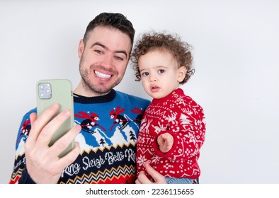 Man holding his beautiful little son with curly hair wearing red Christmas knitted sweater against white background holding smartphone making video call greeting holidays and watching Christmas videos - Shutterstock ID 2236115655