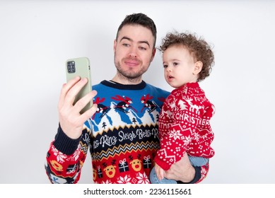 Man holding his beautiful little son with curly hair wearing red Christmas knitted sweater against white background holding smartphone making video call greeting holidays and watching Christmas videos - Shutterstock ID 2236115661