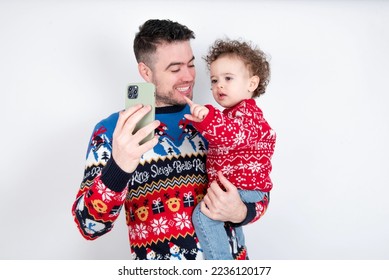 Man holding his beautiful little son with curly hair wearing red Christmas knitted sweater against white background holding smartphone making video call greeting holidays and watching Christmas videos - Shutterstock ID 2236120177