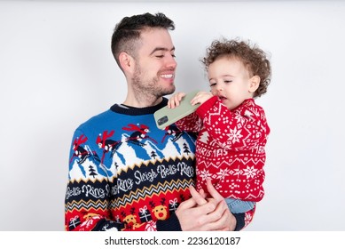 Man holding his beautiful little son with curly hair wearing red Christmas knitted sweater against white background holding smartphone making video call greeting holidays and watching Christmas videos - Shutterstock ID 2236120187