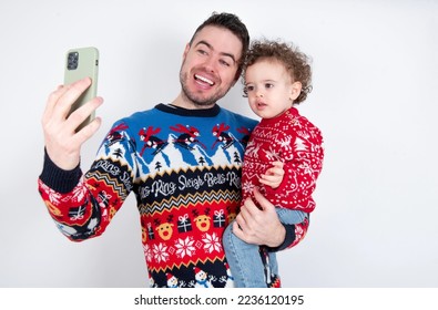 Man holding his beautiful little son with curly hair wearing red Christmas knitted sweater against white background holding smartphone making video call greeting holidays and watching Christmas videos - Shutterstock ID 2236120195