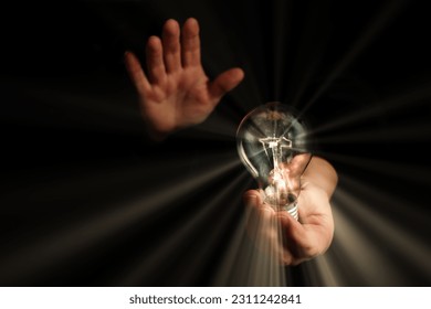 man holding in his hands a lighted light bulb with rays of light on a black background - Shutterstock ID 2311242841